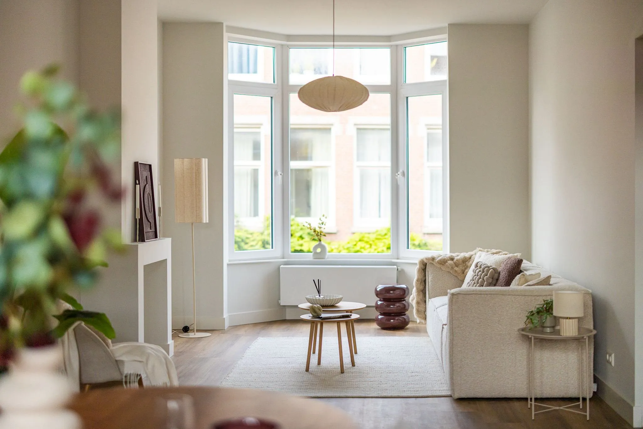 Lichte woonkamer met erker, een witte bouclé bank, ronde houten bijzettafels en een linnen hanglamp
