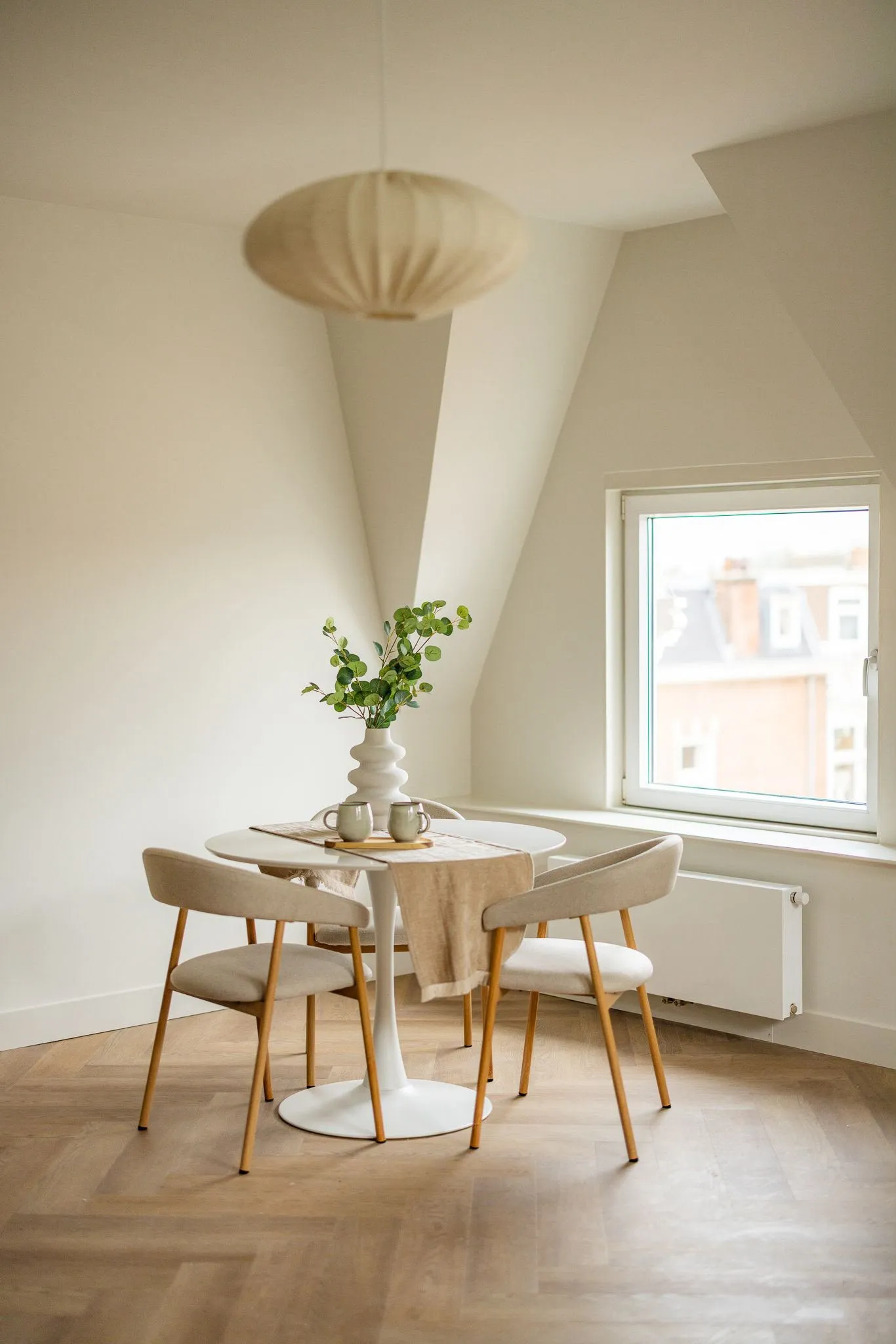 Ronde witte eettafel met beige stoelen op een visgraatvloer, onder een linnen hanglamp bij het raam